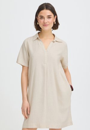 Mladá žena s krátkymi tmavými vlasmi má na sebe béžové šaty s krátkym rukávom a golierom, zlaté kruhové náušnice a drží ruku v vrecku šiat.