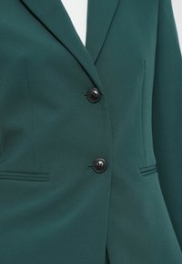 Zelené sako vyrobené z hladkého materiálu, s dvěma černými knoflíky, přizpůsobeným střihem, klopami a bočními kapsami se subtilními detaily prošití.