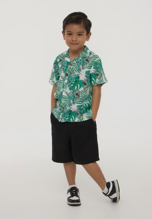 Camicia a maniche corte con stampa di dinosauro verde e foglie, abbinata a pantaloni corti neri. Indossata con sneakers bianche e nere.