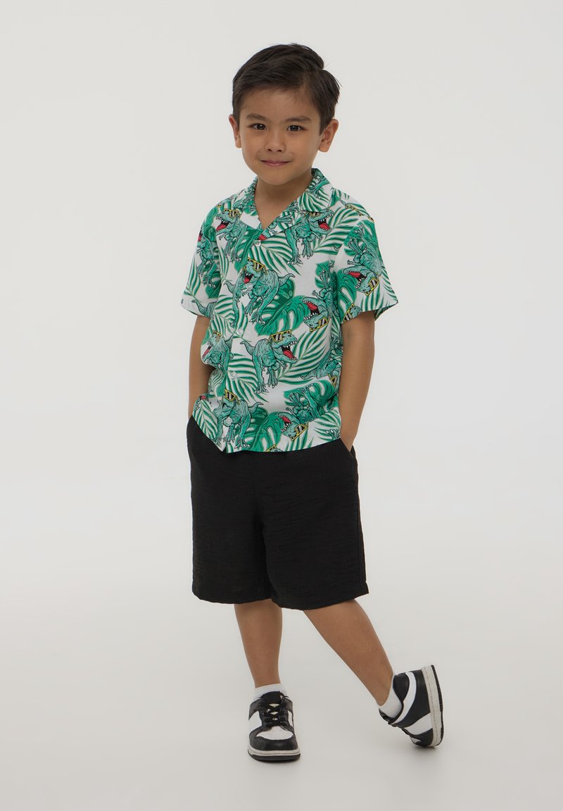Camicia a maniche corte con stampa di dinosauro verde e foglie, abbinata a pantaloni corti neri. Indossata con sneakers bianche e nere.