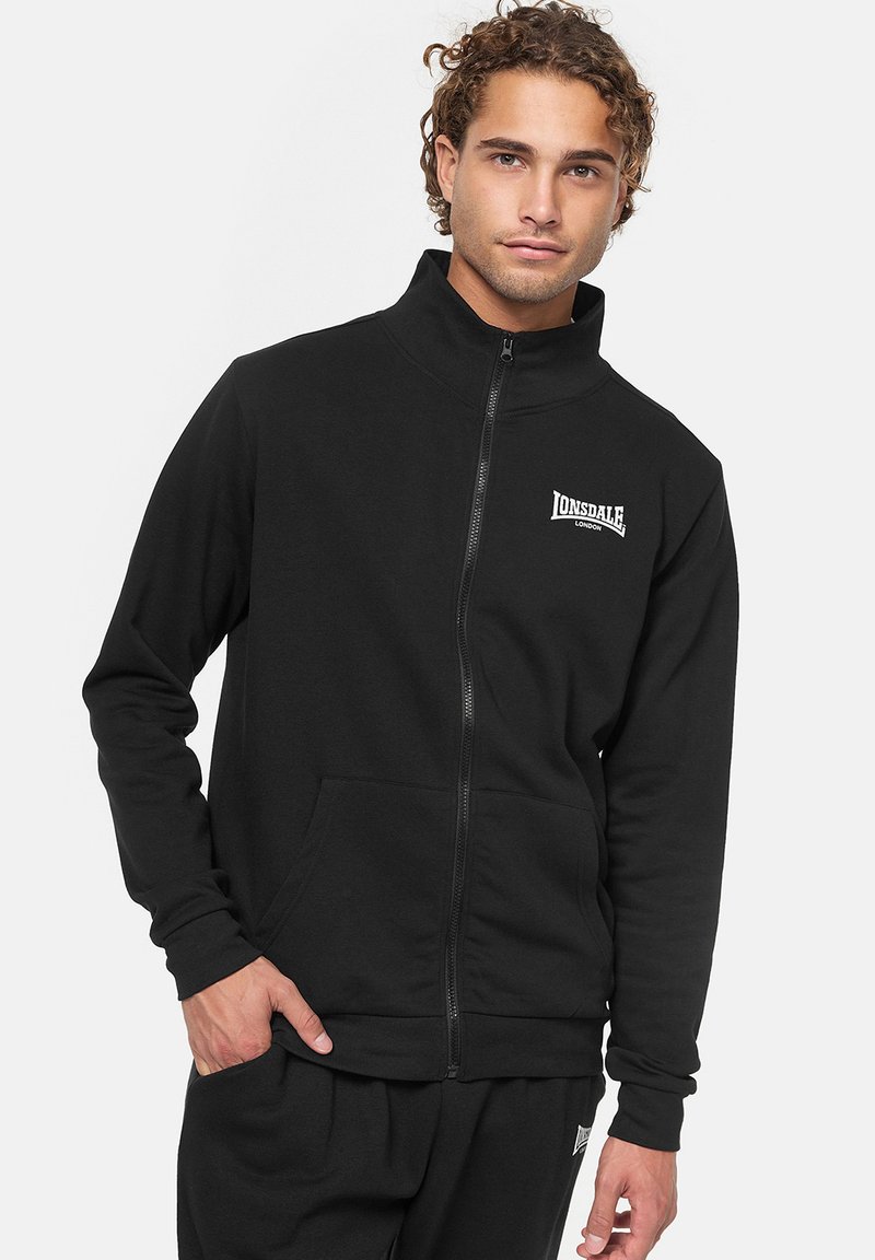 Sudadera negra con cremallera y cuello alto, bolsillos frontales y logo de Lonsdale en el pecho. Fabricada con un tejido suave y texturizado.
