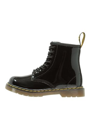 Bota de tobillo negra de cuero brillante con cordones, costuras amarillas y suela de goma marrón translúcida, vista de lado.