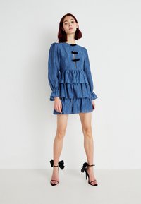 Vestido azul em camadas com tecido texturizado, mangas longas e destacados em botão falso. Sapatos de salto preto com tiras no tornozelo e decoração em forma de laço.