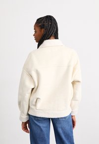 Veste en polaire de couleur crème avec un tissu doux et texturé, dotée d'un col et d'une coupe décontractée, assortie à un jean bleu.