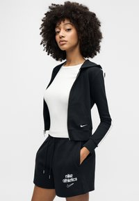 Μαύρο φούτερ με φερμουάρ και λευκή ρίγα στα μανίκια, συνδυασμένο με λευκό εφαρμοστό μπλουζάκι και μαύρο αθλητικό σορτς με λογότυπο και κορδόνι.