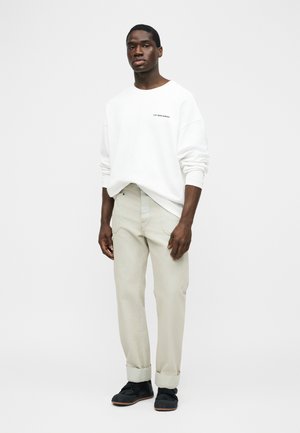 Man draagt een witte sweatshirt met lange mouwen met de tekst "LES BENJAMINS", beige opgerolde broek en zwarte sneakers, staand tegen een witte achtergrond.