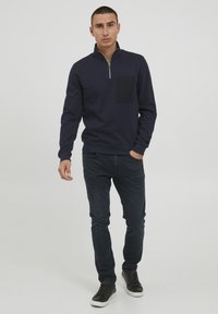 Pulover bleumarin cu guler cu fermoar și buzunar pe piept negru, având o textură solidă. Asortat cu blugi închis la culoare și adidași negri.