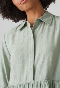 Helles grünes Hemdkleid aus strukturiertem Stoff, mit einem Kragen, Knopfverschluss und Raffung in der Taille.