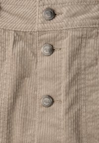 Tissu en velours côtelé beige avec des côtes verticales, présentant trois boutons en argent gravés et une petite boucle en tissu au centre.