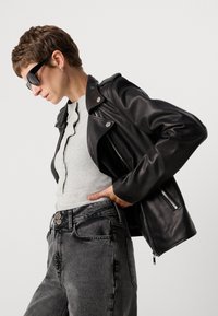 Čierna kožená moto bunda so striebornými prvkami, v kombinácii so svetlošedým rebrovaným topom a šedými džínsami. Modelka pózuje z boku.
