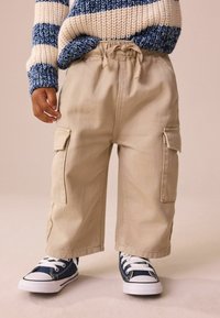 Beige krāsu bikses ar somu kabatām, elastīgu jostu un auklu. Saskaņotas ar tumši ziliem auduma sporta apaviem un baltiem šņorēm.