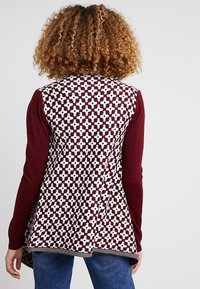 Burgundy kofta med vitt geometriskt mönster, långa ärmar och utsvängd fåll. Mjuk textur med en kontrasterande svart och vit rand längst ner.