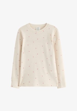 Ung pige stående barfodet iført cremefarvet langærmet top og leggings med mønster af små lyserøde hjerter på hvid baggrund.