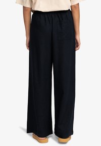 Szerokie spodnie w ciemnym kolorze, wykonane z miękkiego materiału, z elastycznym paskiem w talii i tylną kieszenią. Projekt charakteryzuje się luźnym krojem oraz minimalnymi detalami.
