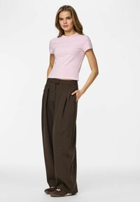 T-shirt à manches courtes rose pâle associée à un pantalon taille haute, jambes larges, de couleur marron foncé, avec plis sur le devant et poches latérales. Baskets marron.