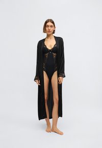 Μακρύ μαύρο σατέν ρόμπα με δαντελένιες λεπτομέρειες, που φοριέται πάνω από μαύρο δαντελένιο μαγιό. Διαθέτει μακριά μανίκια και ανοιχτό μπροστινό μέρος. Ομαλή υφή, κομψός σχεδιασμός.