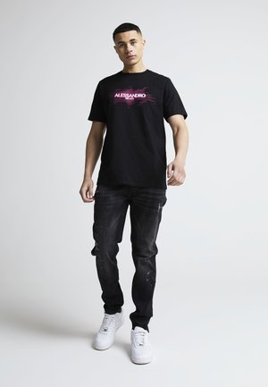 Ung mand iført sort Alessandro Milano T-shirt, mørke slidte jeans og hvide sneakers, stående mod en ensartet lys baggrund.