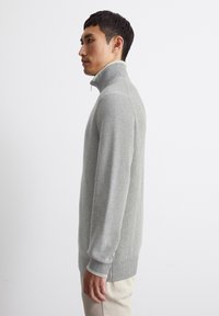Light gray ribbed sweater with a high collar, featuring a zipper and fitted sleeves, worn by a model against a white background.