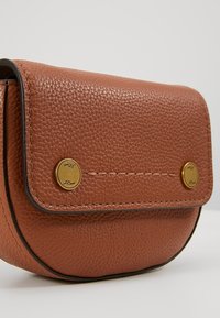 Petit sac bandoulière en cuir brun texturé avec rabat et deux fermoirs boutons dorés sur un fond uni.