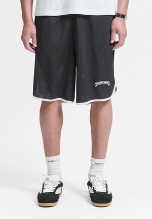 Persoon die zwarte mesh shorts met witte biezen draagt, witte sokken met de tekst "RETERNITY", zwart-witte sneakers en een polshorloge.