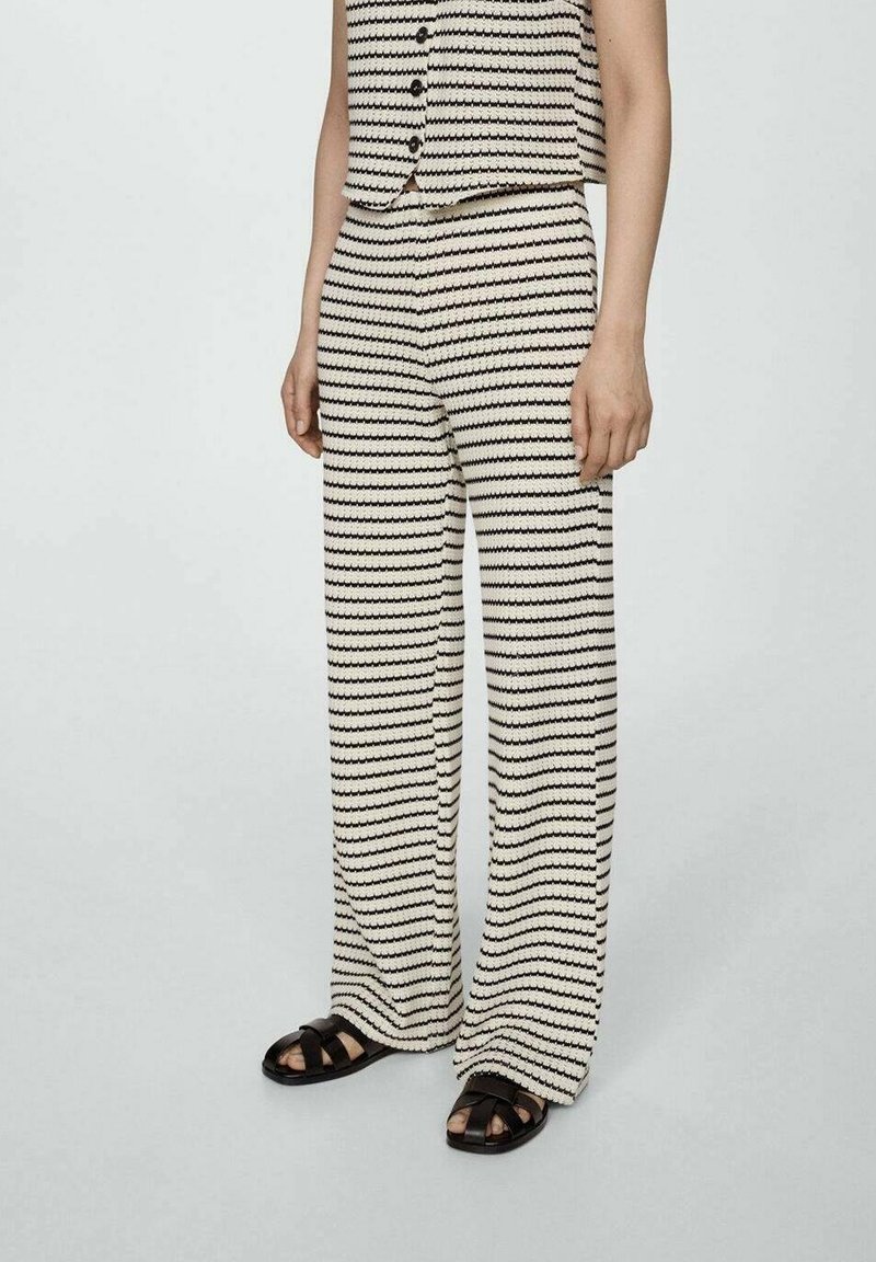 Osoba v černobílých pruhovaných širokých kalhotách a odpovídajícím bezramenním topu s černými sandály, stojící na jednobarevném pozadí.