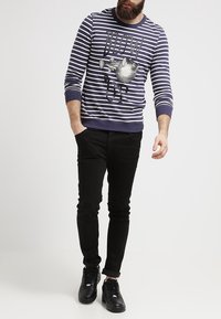 Sudadera a rayas en azul marino y blanco con un estampado gráfico y texto, combinada con jeans negros ajustados y zapatillas negras.