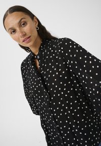 Black button-up shirt with white polka dots. Lightweight fabric, ruffled collar, and loose sleeves. Features a front button closure.