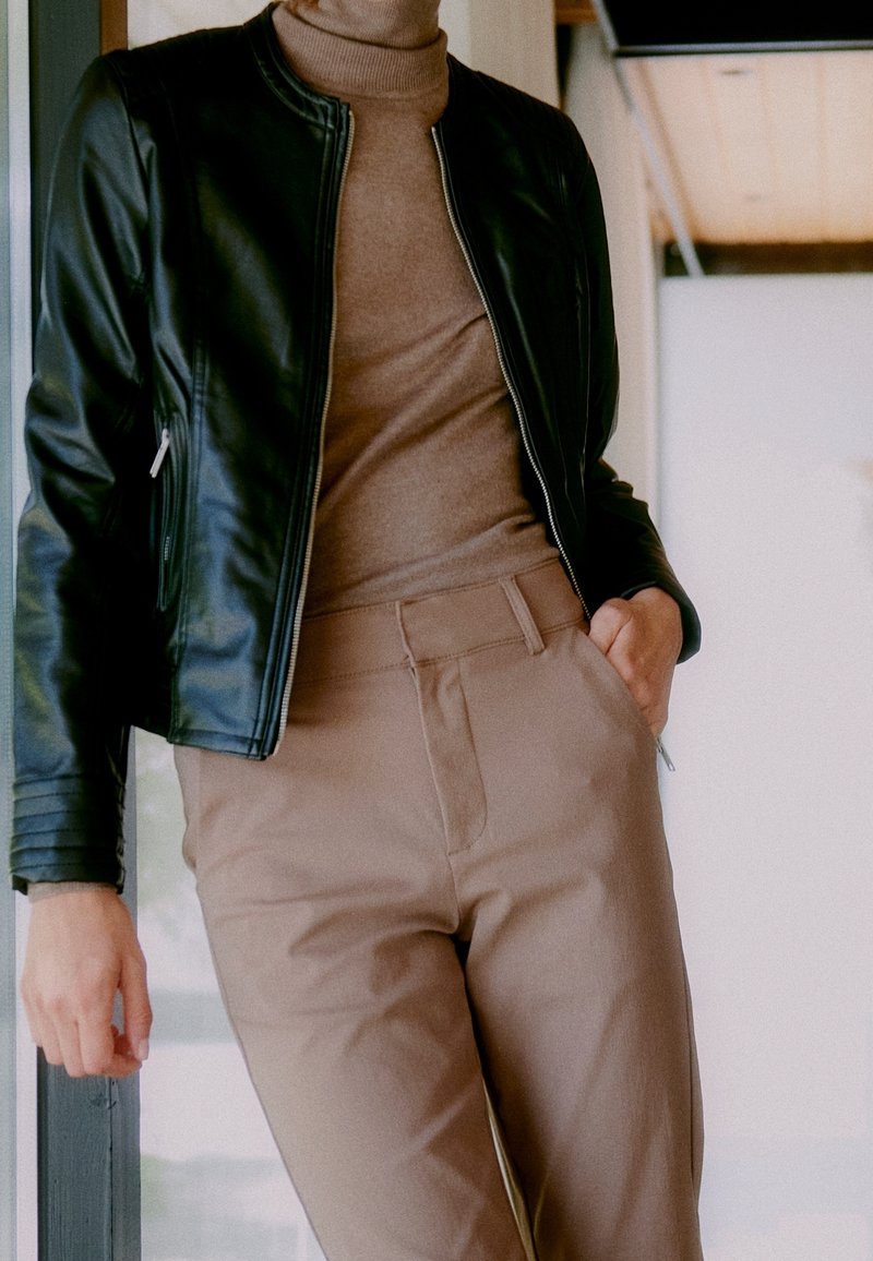 Veste en cuir noire à la coupe ajustée, avec des poignets côtelés, portée par-dessus un pull à col roulé marron clair et un pantalon beige, avec des poches latérales.