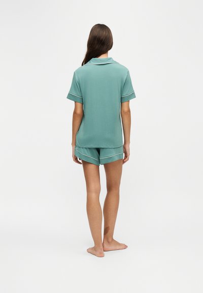 Femme debout pieds nus portant un haut vert à manches courtes et un short assorti avec un bord blanc, vue de dos sur fond blanc.