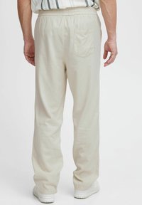 Hellbeige Hosen mit elastischem Bund, einer hinteren Tasche und lockerer Passform, kombiniert mit weißen Turnschuhen vor einem neutralen Hintergrund.