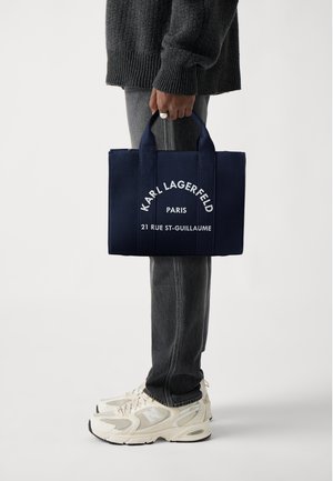 Osoba w ciemnym swetrze i dżinsach trzyma granatową torbę typu tote marki Karl Lagerfeld z białym napisem, nosi białe sneakersy New Balance.