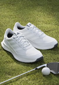 White athletic shoes with textured uppers and lacing, featuring three side stripes. Positioned on grass beside a golf club and ball.