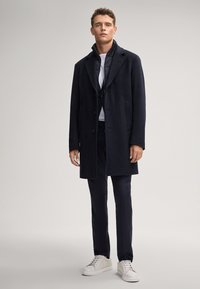 Cappotto di lana scuro con ampi risvolti, tre bottoni, due tasche frontali, abbinato a una t-shirt bianca, pantaloni scuri e sneakers bianche.