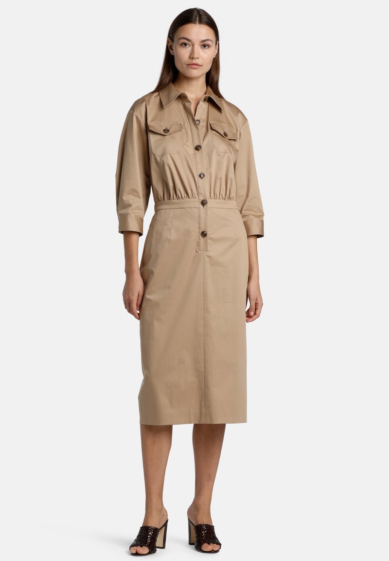 Tan button-up dress with a fitted waist and two chest pockets. Three-quarter rolled sleeves, knee-length hem, and smooth texture. Brown buttons.