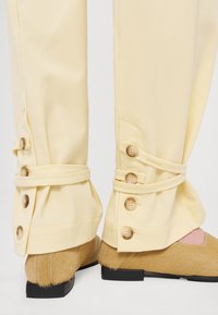 Gambe inferiori con pantaloni beige dotati di cinturini con bottoni sui polsini, abbinati a scarpe slip-on color cuoio con texture e suole nere.