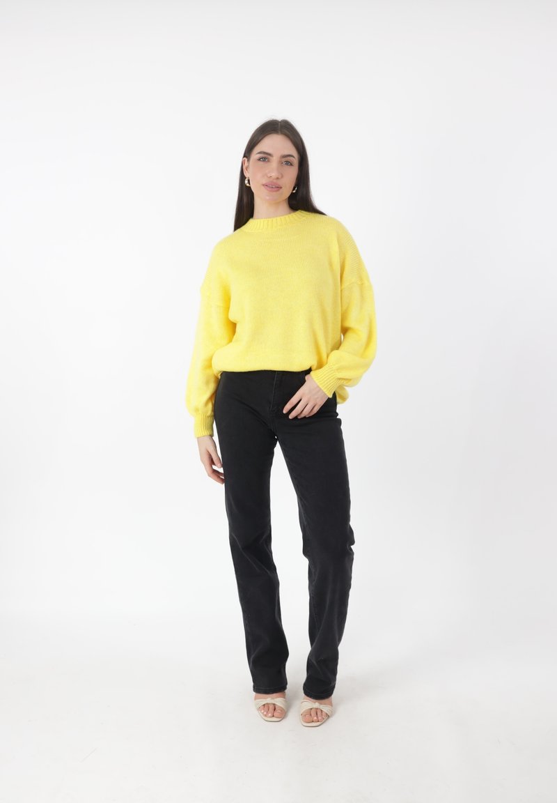 Maglione giallo a maglia con collo alto e spalle scese, abbinato a pantaloni neri aderenti e scarpe con punta aperta color nude. Tessuto liscio.