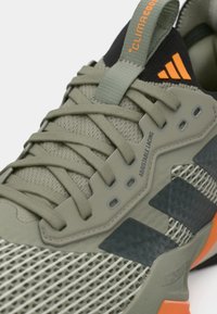 Αθλητικό παπούτσι σε ελιά πράσινο με πλεκτό επάνω μέρος, ρυθμιζόμενο σύστημα κορδονιών, μαύρες λεπτομέρειες και πορτοκαλί λογότυπα. Διαθέτει υλικό με υφή.