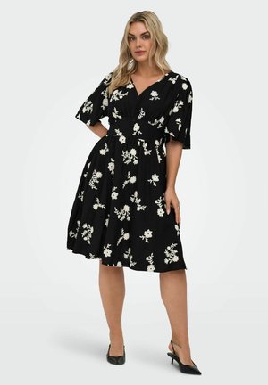 Kvinde iført en sort knælængde kjole med hvide blomsterprint og flagrende ærmer, stående med den ene hånd på hoften og sorte hæle.