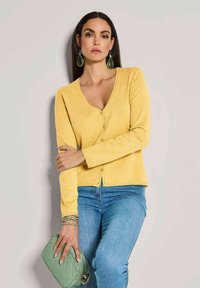 Gelbe Strickjacke mit V-Ausschnitt, langen Ärmeln und durchgehender Knopfleiste. Kombiniert mit blauen Jeans und einer grünen, gesteppten Handtasche.