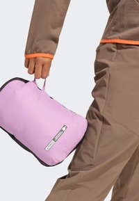 Packbar rosa isoleringsväska med svarta nätdetaljer, hålls i handen. Brun jacka med orangea detaljer vid ärmsluten.