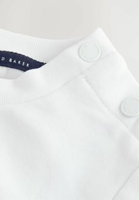 Weißes Baumwollkleidungsstück mit geripptem Rundhalsausschnitt, versehen mit zwei dekorativen weißen Knöpfen auf der Schulter und einem marineblauen Etikett innen.