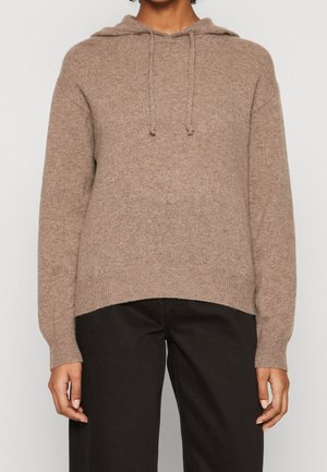 Personne portant un sweat à capuche taupe en tricot avec cordons, associé à un pantalon noir ample, visible du cou aux hanches.