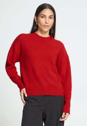 Femme aux longs cheveux foncés portant un pull en maille rouge et un pantalon noir, debout avec une main sur la hanche devant un fond uni.