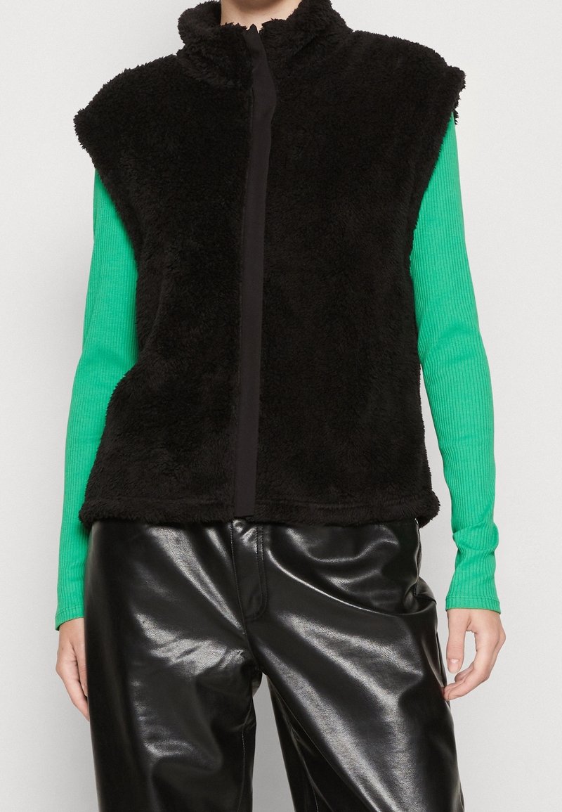 Veste sans manches en polaire noire avec un col montant et une fermeture éclair, portée sur un haut texturé vert à manches longues, associée à un pantalon noir brillant.