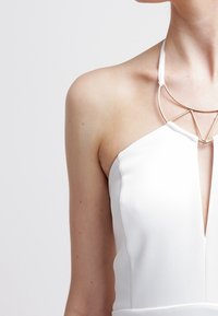 Bílé šaty s halter krkem a hlubokým výstřihem do V, s trojúhelníkovým stříbrným akcentem na límci. Hladká tkanina s minimální texturou.