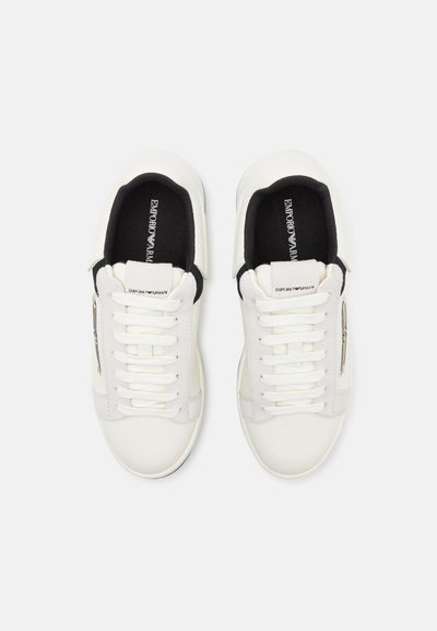 Sneakers bianchi con superficie in pelle liscia, fodera interna nera, lacci piatti e un logo sottile sul tallone. Design semplice, stile a taglio basso.