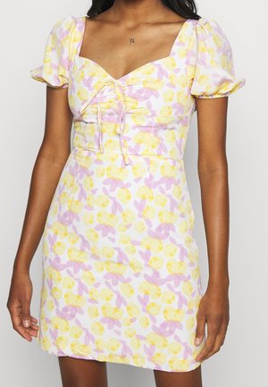 Nainen pukeutuneena lyhyeen, vartaloa myötäilevään mekkoon, jossa on pussihihat, sydämenmuotoinen pääntie ja valkoisella kankaalla keltainen ja pinkki kukkakuvio.