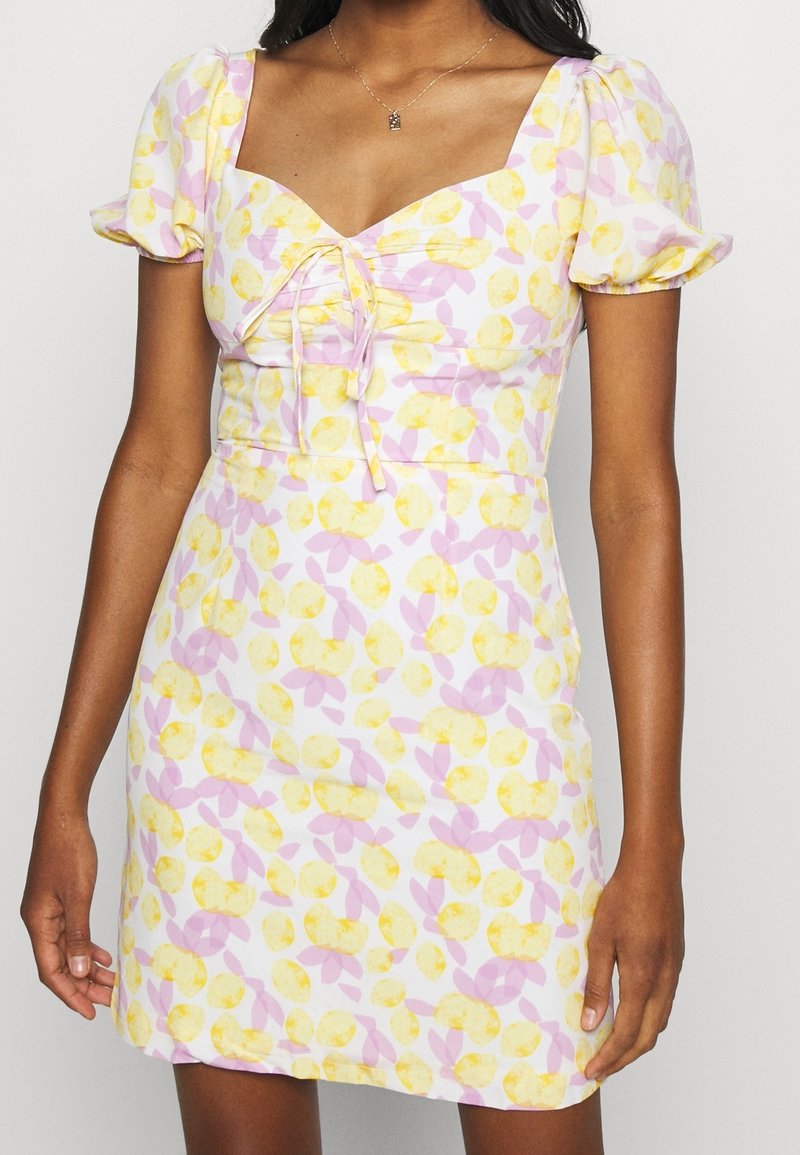 Femme portant une robe courte et ajustée avec des manches bouffantes, un décolleté en cœur, et un motif floral jaune et rose sur tissu blanc.