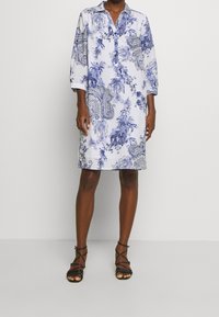 Donna che indossa un abito bianco lungo fino al ginocchio con motivi floreali e paisley blu e sandali neri con tacco alto e bretelle.