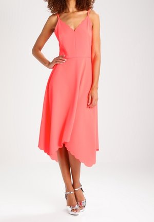 Kobieta w koralowo-różowej sukience midi bez rękawów z falistym dekoltem i dołem, w połączeniu z srebrnymi sandałami na wysokim obcasie.